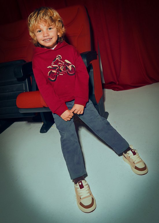 Jongetje zit in een bioscoopstoel met een bordeauxrode hoodie met motorprint en grijze broek.