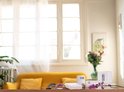 Twee witte Netatmo slimme thermostaten met digitale display op een houten tafel, met bloemen en een gele bank op de achtergrond.