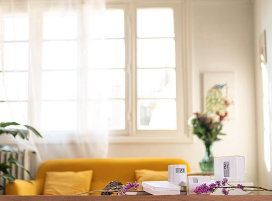 Twee witte Netatmo slimme thermostaten met digitale display op een houten tafel, met bloemen en een gele bank op de achtergrond.