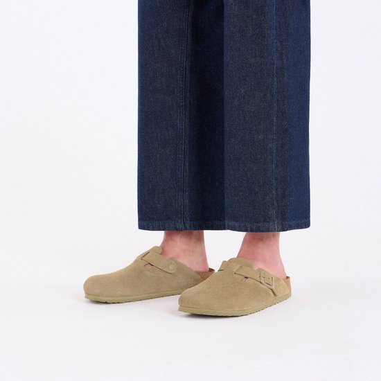 Voeten in spijkerbroek en beige suède Birkenstock Boston slippers, bekeken vanaf de voorkant.
