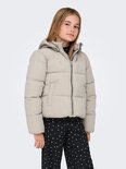 Meisje met blonde staartjes draagt een lichtbeige pufferjack met capuchon, handen in de zakken.