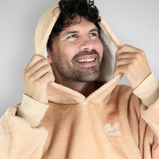 Een man met krullend bruin haar trekt de capuchon van een beige hoodie deken omhoog. Hij lacht en kijkt naar rechts.