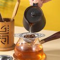 Zilverkleurige theezeef met gaasfilter en versierde vleugel handvatten, boven een glazen theepot waaruit heet water wordt gegoten.