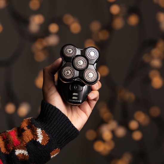 Een hand met kersttrui houdt een zwart Remington hoofdscheerapparaat vast met lichten op de achtergrond.