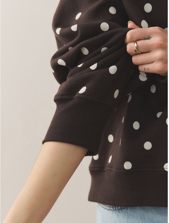 Close-up van de mouw van een donkerbruine sweater met witte polkadotprint, een hand trekt de mouw op.