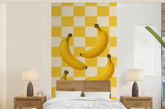 Fotobehang met gele bananen op een geel-wit geblokt patroon, boven een houten hoofdeinde. Een bed met wit beddengoed.