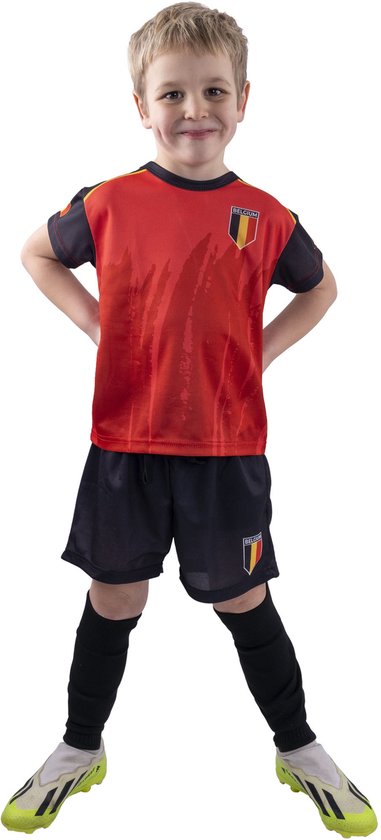 Jongetje met een glimlach in voetbaltenue, rood shirt met Belgische vlag, zwarte broek en handen in de zij.