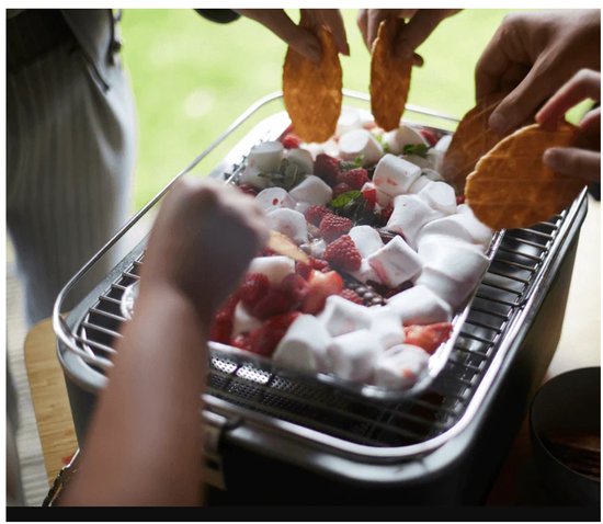 Mensen maken s'mores boven een tafelbarbecue met aardbeien, marshmallows en koekjes.
