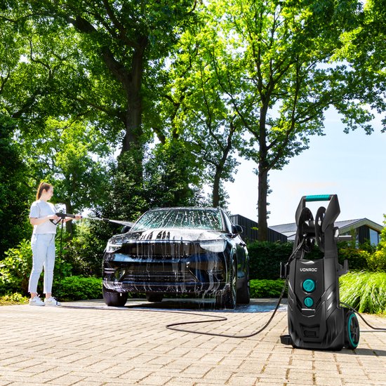 Vrouw met hogedrukreiniger bij een zwarte auto, omgeven door groen.