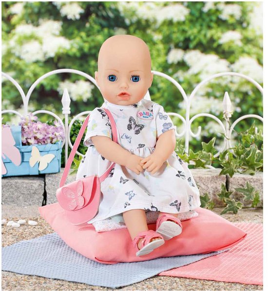 Baby Born pop in witte jurk met vlinderprint, roze tasje en roze schoentjes. Zit op roze kussen.