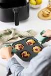 Handen nemen een donkergrijze muffinvorm met 4 bessenmuffins uit een heteluchtfriteuse met lichtgroene grepen.