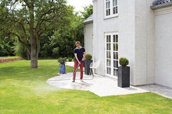 Man spuit met een blauwe Nilfisk hogedrukreiniger een stenen terras schoon in een tuin met groen gras en een huis.