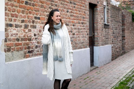 Een vrouw draagt een gebreide sjaal in grijs gemêleerd met een geleidelijke overgang van licht naar donkergrijs. Ze draagt ook een gebreide vest en jurk.