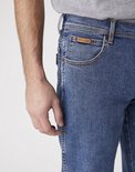 Close-up van de voorkant van een spijkerbroek met een bruin Wrangler-label op de kleine zak, een man met grijze t-shirt.