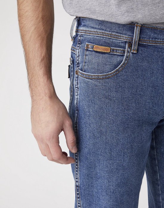 Close-up van de voorkant van een spijkerbroek met een bruin Wrangler-label op de kleine zak, een man met grijze t-shirt.