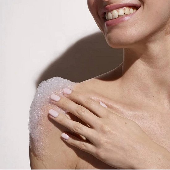 Een vrouw met lichte nagellak lacht, met haar schouder vol schuim en zeepbellen.