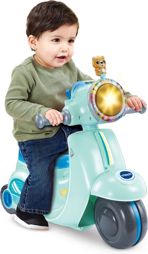 Jongen met donker haar, groene trui en spijkerbroek zit op een lichtgroene driewielige scooter met blauw zadel.