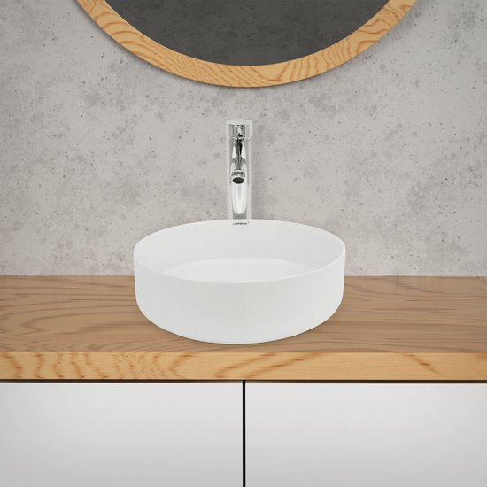 Ronde witte wastafel op een houten meubel onder een ronde spiegel met betonnen achterwand.