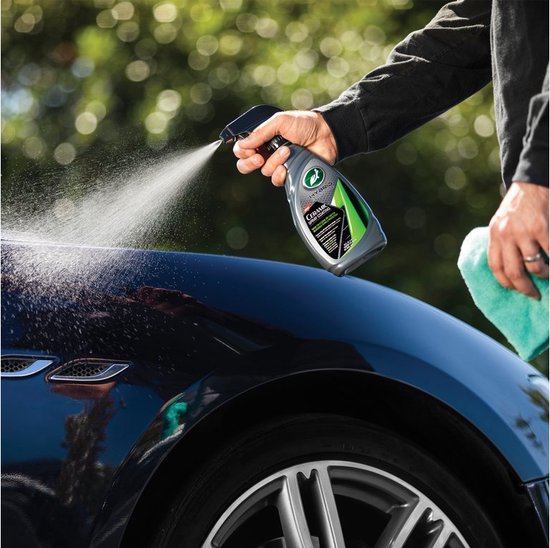 Main vaporisant le flacon gris aux accents verts et noirs sur le capot d'une voiture foncée, avec un chiffon vert à côté.