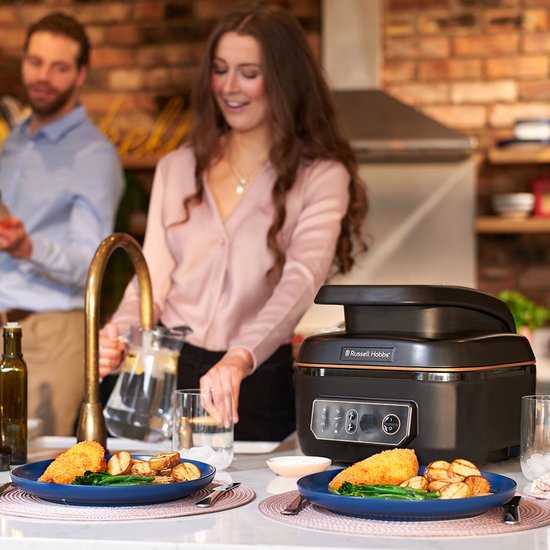 Russell Hobbs airfryer op aanrecht, met gegrilde vis en aardappelen op borden, en een stel in de achtergrond.
