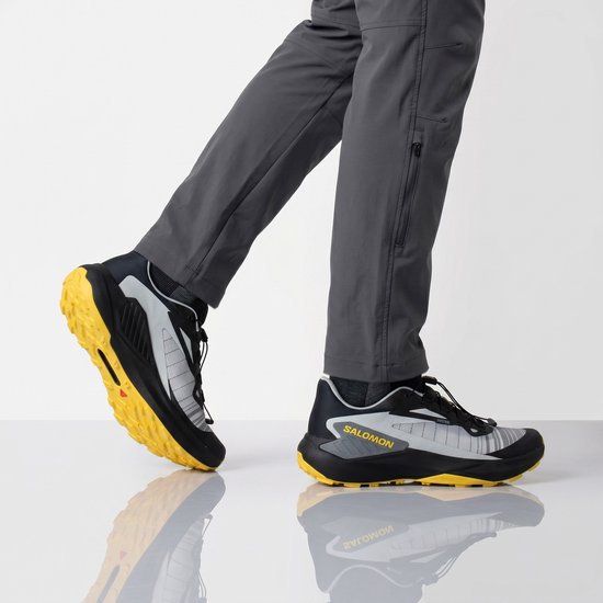 Model in grijze Salomon trailrunningschoenen met gele zolen en donkergrijze broek.