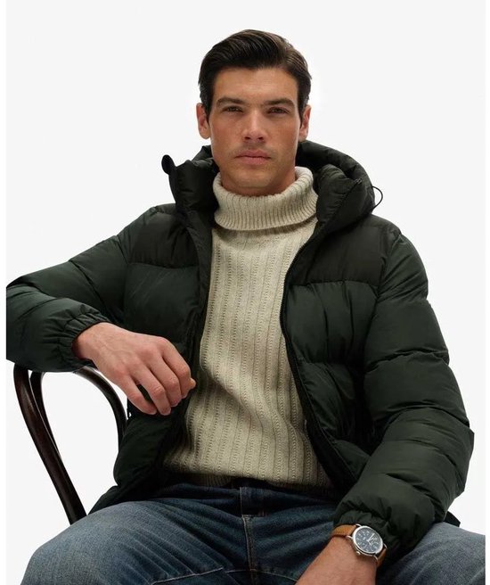 Man met beige coltrui en donkergroene jas met capuchon, zittend.