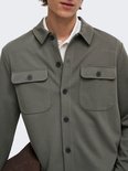 Mannelijk model met olijfgroene, lange mouwen oversized shirt, witte polo eronder en draagt een bruine tas.
