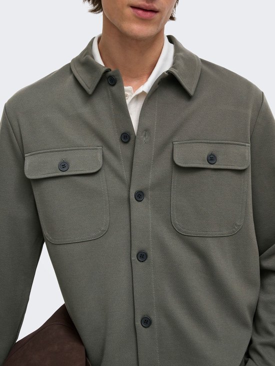 Mannelijk model met olijfgroene, lange mouwen oversized shirt, witte polo eronder en draagt een bruine tas.