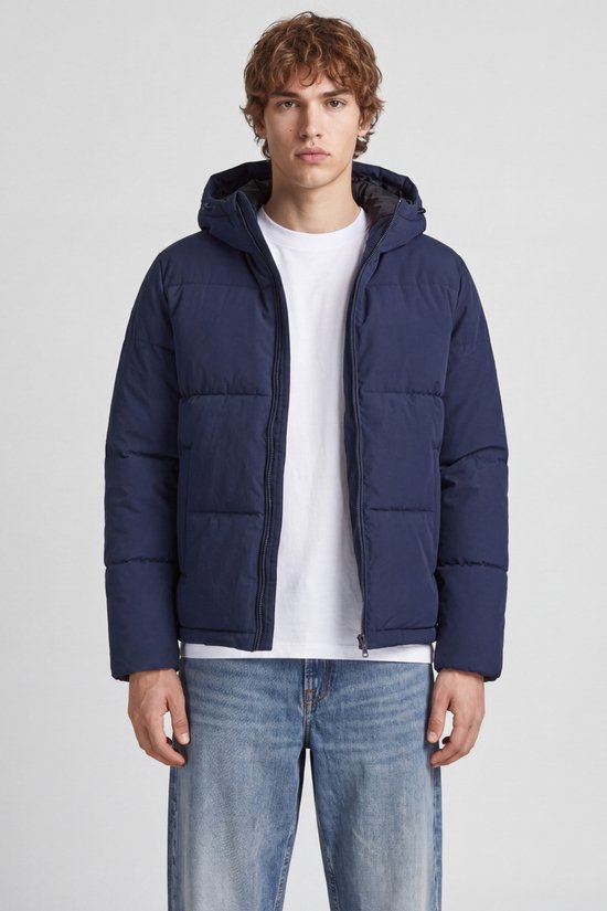 Man met donkerblauwe pufferjas met capuchon, witte t-shirt en wijde spijkerbroek.