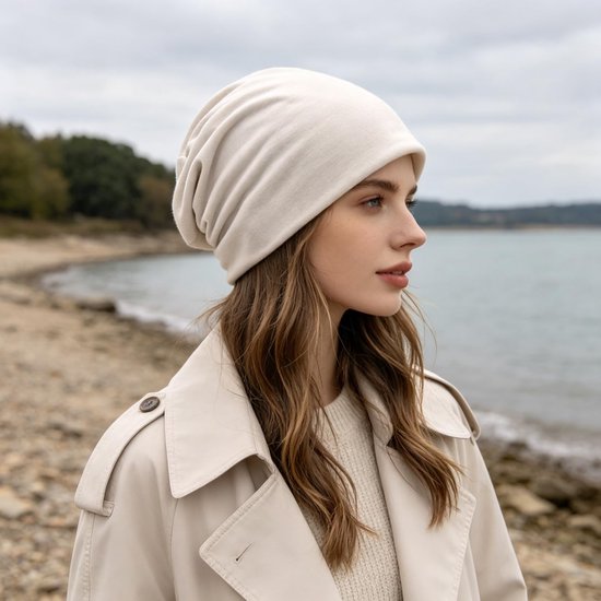 Model met een beige katoenen beanie en trenchcoat, staand aan de waterkant.
