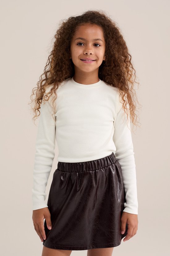 Meisje met krullend bruin haar, draagt crèmekleurig T-shirt van ribstof en een bruine rok.