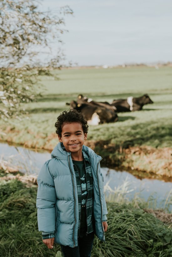 Jongen in een lichtblauwe pufferjas met geruite blouse, lachend in een landelijke omgeving.