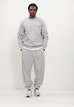 Man staand in grijze New Balance sweater met wit logo en bijpassende joggingbroek.