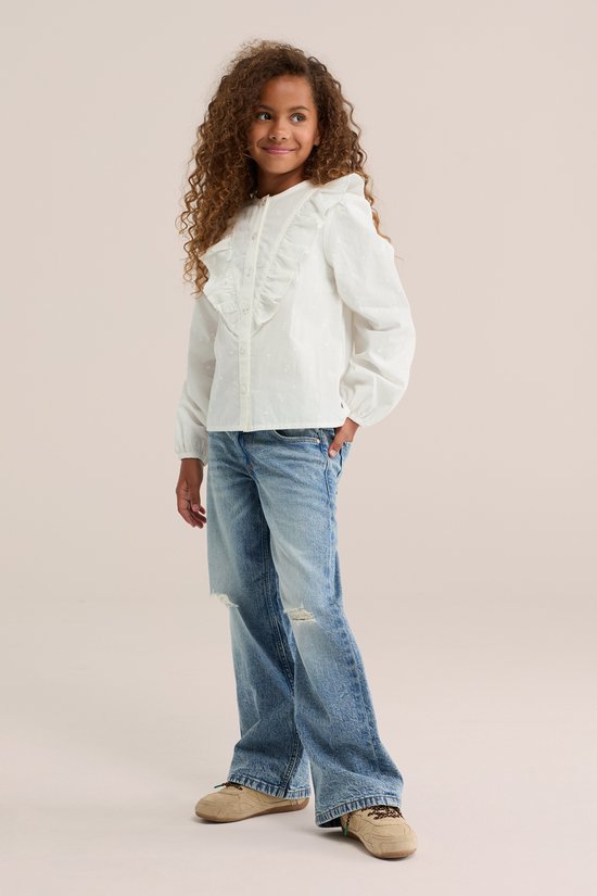 Meisje met krullend haar staat met de hand in de zak van haar spijkerbroek, ze draagt een witte blouse met ruches en geborduurde bloemetjes.