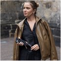 Vrouw met bruine jas en zwart shirt, vasthoudend aan een zwart telefoonhoesje met een gevlochten koord met hartjes.