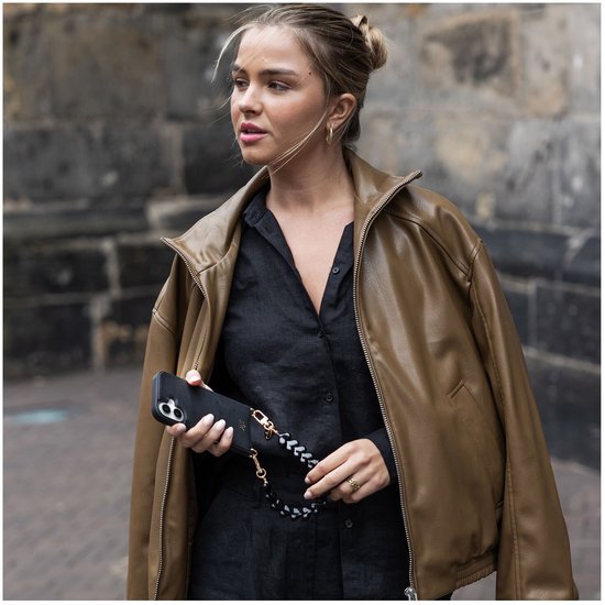 Vrouw met bruine jas en zwart shirt, vasthoudend aan een zwart telefoonhoesje met een gevlochten koord met hartjes.