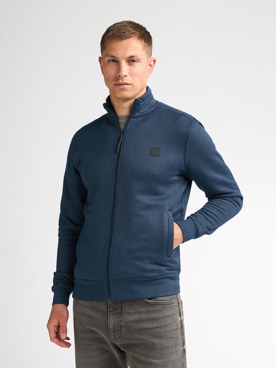 Model in een blauw vest met ritssluiting, legergroen T-shirt en grijze jeans. Handen in de zakken.