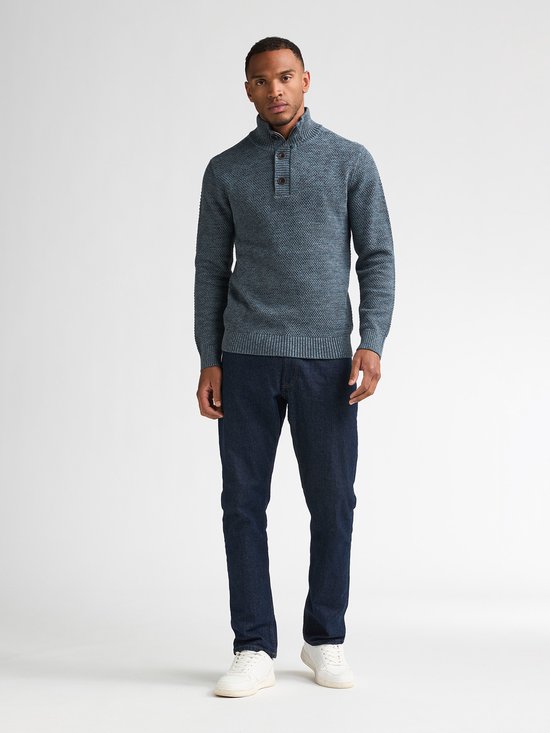 Man in blauwe gemêleerde sweater met hoge kraag, donkerblauwe jeans en witte sneakers.
