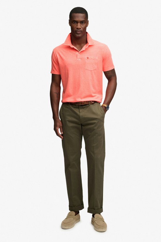 Man in oranje polo met opgestoken kraag, broek en beige loafers. Staat bij een stoel.