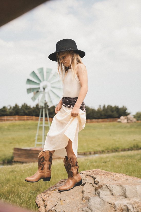 Meisje met blonde haren en zwarte hoed staat op een steen, draagt een lichte jurk en cognac cowboylaarzen.