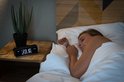 Vrouw slaapt naast een zwarte Lenco wekkerradio met tijd '23:40' en zender 'BBC WORLD SERVICE' op het nachtkastje.