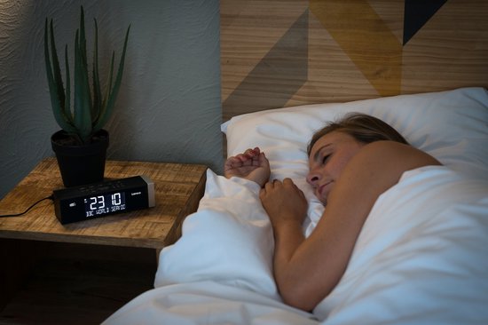 Vrouw slaapt naast een zwarte Lenco wekkerradio met tijd '23:40' en zender 'BBC WORLD SERVICE' op het nachtkastje.