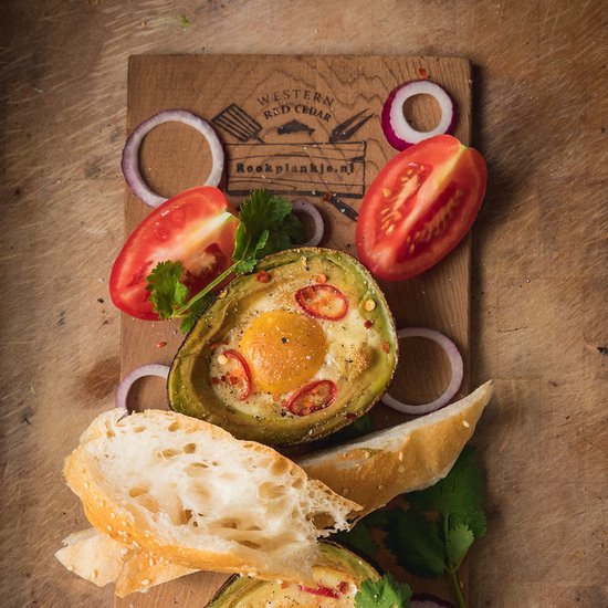 Gevulde avocado met ei, tomaat, rode ui en brood op een cederhouten plank.