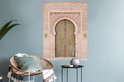 Une porte marocaine couleur sable avec des détails dorés et une sculpture sur pierre complexe, accrochée au-dessus d'une chaise en rotin et d'une table d'appoint.