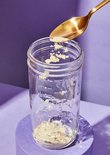Een gouden lepel voegt lichtgeel poeder toe aan een lege glazen pot.