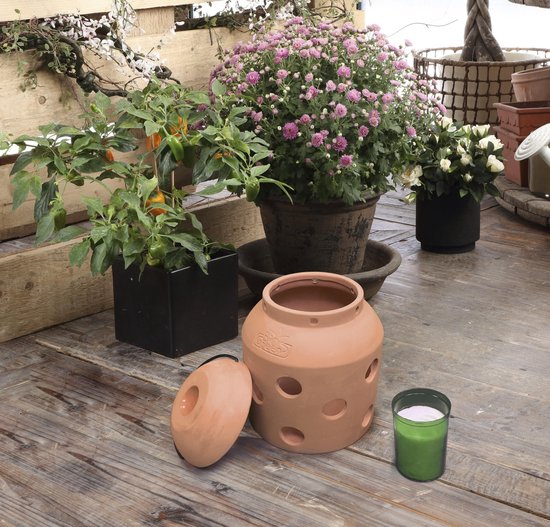 Terracotta kaarsenwarmer met deksel, naast een groene kaars en planten op een houten tafel.