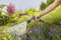 Hand met GARDENA Comfort EcoPulse spuitpistool, sproeit water op bloemen in de tuin.