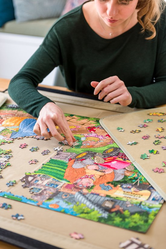 Een vrouw met een groene trui legt een puzzel met kabouters en een tractor. Stukjes liggen verspreid op de mat.