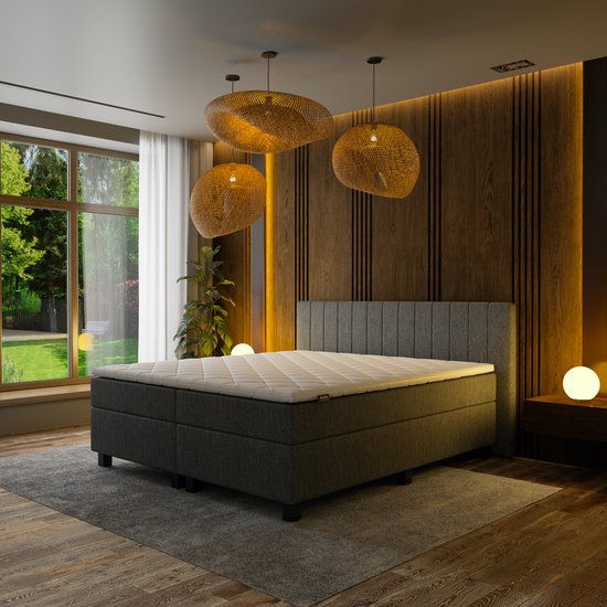 Grijze boxspring bed met wit topmatras, in een slaapkamer met houten lamellenwand en grote ramen naar de tuin.