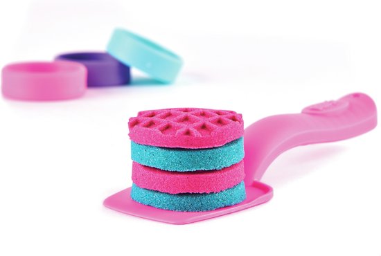 Roze en turquoise gestapelde kinetische zand schijven op een roze spatel, met gekleurde ringen op de achtergrond.